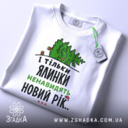 Футболка ялинки новий рік, біла, складена на поверхні, фон світлий, принт з ялинкою та текстом у зеленому та чорному кольорах, з біркою на рукаві.