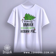 Футболка ялинки новий рік, біла, підвішена на вішалці, фон світлий, принт з ялинкою та текстом у зеленому та чорному кольорах.