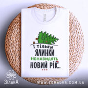 Футболка ялинки новий рік, біла, складена на плетеній кошику, фон нейтральний, принт з ялинкою та текстом у зеленому та чорному кольорах.