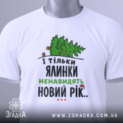 Футболка ялинки новий рік, біла, складена на поверхні, фон світлий, принт з ялинкою та текстом у зеленому та чорному кольорах, з біркою на рукаві.