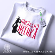 Футболка ідеальна жінка, біла, зображена в складеному вигляді, фон світло-сірий, принт з силуетом відьми та написом червоним шрифтом 'ідеальна жінка', з етикеткою на комірці.