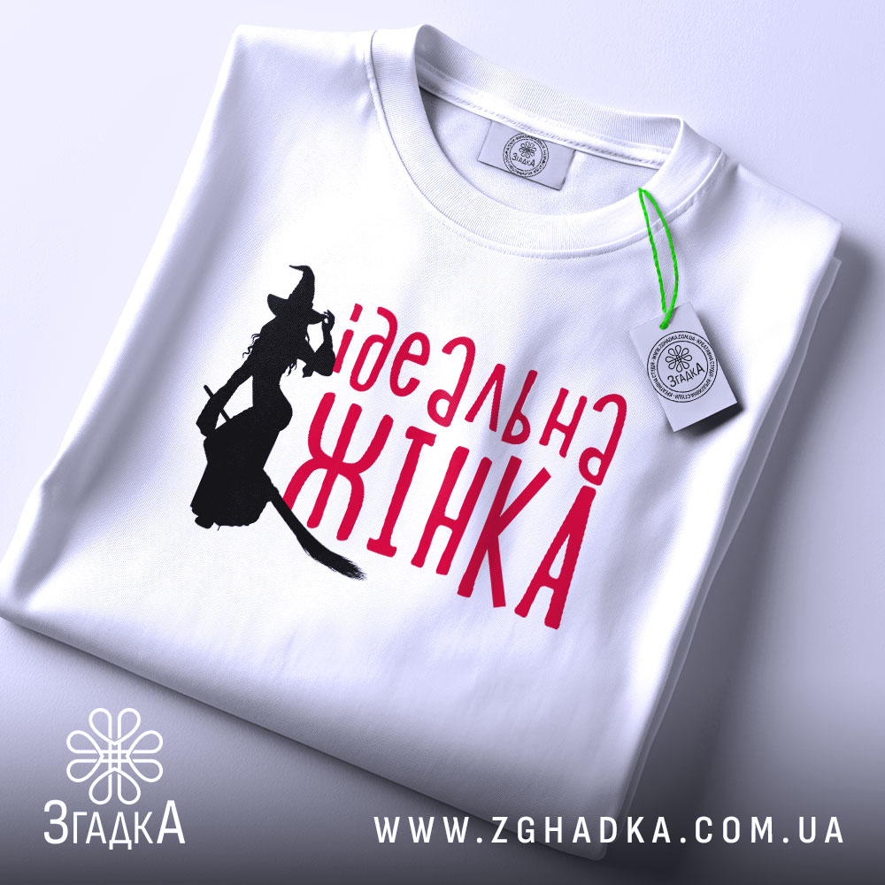 Футболка ідеальна жінка, біла, зображена в складеному вигляді, фон світло-сірий, принт з силуетом відьми та написом червоним шрифтом 'ідеальна жінка', з етикеткою на комірці.
