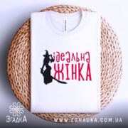 Футболка ідеальна жінка, біла, зображена в складеному вигляді на плетеній основі, фон світло-сірий, принт з силуетом відьми та написом червоним шрифтом 'ідеальна жінка'.