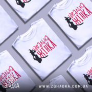 Футболка ідеальна жінка, біла, зображена в розкладеному вигляді, фон світло-сірий, кілька футболок з принтом з силуетом відьми та написом червоним шрифтом 'ідеальна жінка' на фоні.