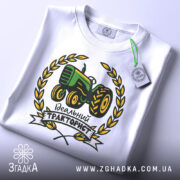 Футболка ідеальний тракторист, біла, складена на столі, фон світло-сірий, принт з трактором зеленого кольору, оточений жовтими листям і написом 'Ідеальний тракторист', на комірці видна етикетка.