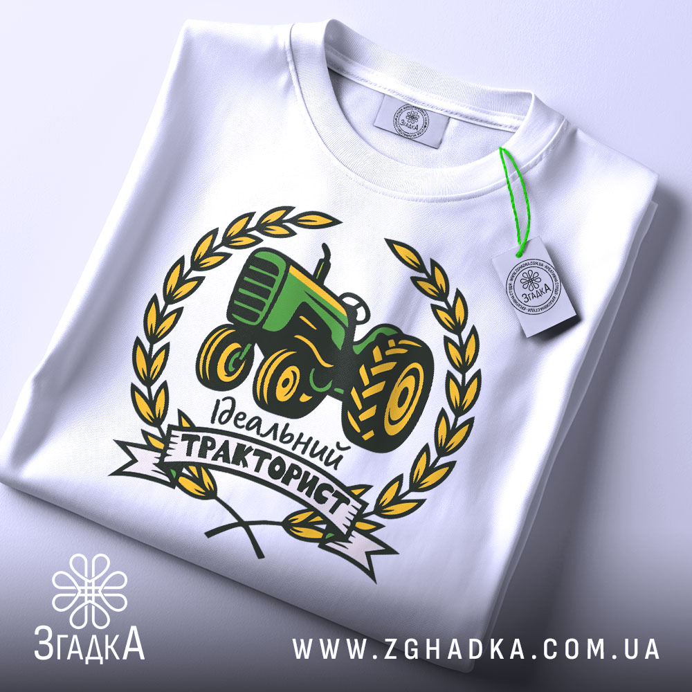 Футболка ідеальний тракторист, біла, складена на столі, фон світло-сірий, принт з трактором зеленого кольору, оточений жовтими листям і написом 'Ідеальний тракторист', на комірці видна етикетка.