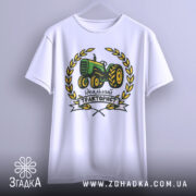 Футболка ідеальний тракторист, біла, підвішена на вішалці, фон світло-сірий, принт з трактором зеленого кольору, оточений жовтими листям і написом 'Ідеальний тракторист'.