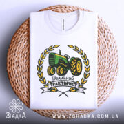 Футболка ідеальний тракторист, біла, складена на плетеній тарілці, фон білий, принт з трактором зеленого кольору, оточений жовтими листям і написом 'Ідеальний тракторист'.