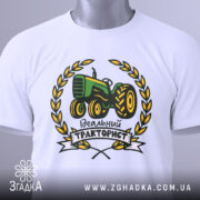 Футболка ідеальний тракторист, біла, складена на столі, фон світло-сірий, принт з трактором зеленого кольору, оточений жовтими листям і написом 'Ідеальний тракторист', на комірці видна етикетка.