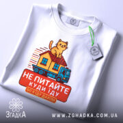 Футболка їду дахом, біла, зображена з верхнього ракурсу, фон світлий, принт з котом на даху будинку, що їде на вантажівці, текст червоним кольором, на комірці видна етикетка.