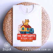 Футболка їду дахом, біла, зображена зверху, фон плетений, принт з котом на даху будинку, що їде на вантажівці, текст червоним кольором.