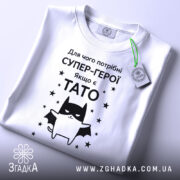 Футболка є тато, білого кольору, зображена в горизонтальному положенні, фон світло-сірий, принт чорного кольору з зображенням кота-супергероя та текстом на українській мові, з етикеткою зеленого кольору.