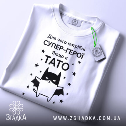 Футболка є тато, білого кольору, зображена в горизонтальному положенні, фон світло-сірий, принт чорного кольору з зображенням кота-супергероя та текстом на українській мові, з етикеткою зеленого кольору.