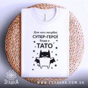 Футболка є тато, білого кольору, зображена в горизонтальному положенні на плетеній основі, фон світло-сірий, принт чорного кольору з зображенням кота-супергероя та текстом на українській мові.