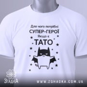 Футболка є тато, білого кольору, зображена в горизонтальному положенні, фон світло-сірий, принт чорного кольору з зображенням кота-супергероя та текстом на українській мові, з етикеткою зеленого кольору.