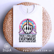 Футболка єдинороги іронія, біла футболка з коротким рукавом, складена на плетеному кошику, фон світло-сірий, принт з чорним малюнком руки з середнім пальцем, оточений кольоровими елементами, включаючи райдугу та єдинорогів.