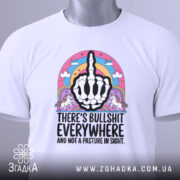 Футболка єдинороги іронія, біла футболка з коротким рукавом, зображена зверху на столі, фон світло-сірий, принт з чорним малюнком руки з середнім пальцем, оточений кольоровими елементами, включаючи райдугу та єдинорогів, на футболці є етикетка.