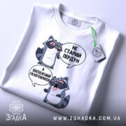 Футболка єнот пердун білого кольору, складена на плоскій поверхні, фон світлий. Принт з двома єнотами в окулярах, текст той же, що й на попередньому фото. На футболці прикріплений зелений ярлик.