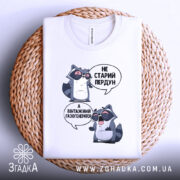 Футболка єнот пердун білого кольору, складена на плоскій поверхні з плетеним елементом на фоні. Принт з двома єнотами в окулярах, текст той же, що й на інших фото.