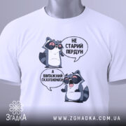 Футболка єнот пердун білого кольору, складена на плоскій поверхні, фон світлий. Принт з двома єнотами в окулярах, текст той же, що й на попередньому фото. На футболці прикріплений зелений ярлик.