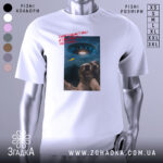 Футболка інопланетяни атакують Землю, біла футболка на манекені, ракурс спереду, фон нейтральний, принт з двома собаками, що сміються, та космічним кораблем на фоні з українським прапором.