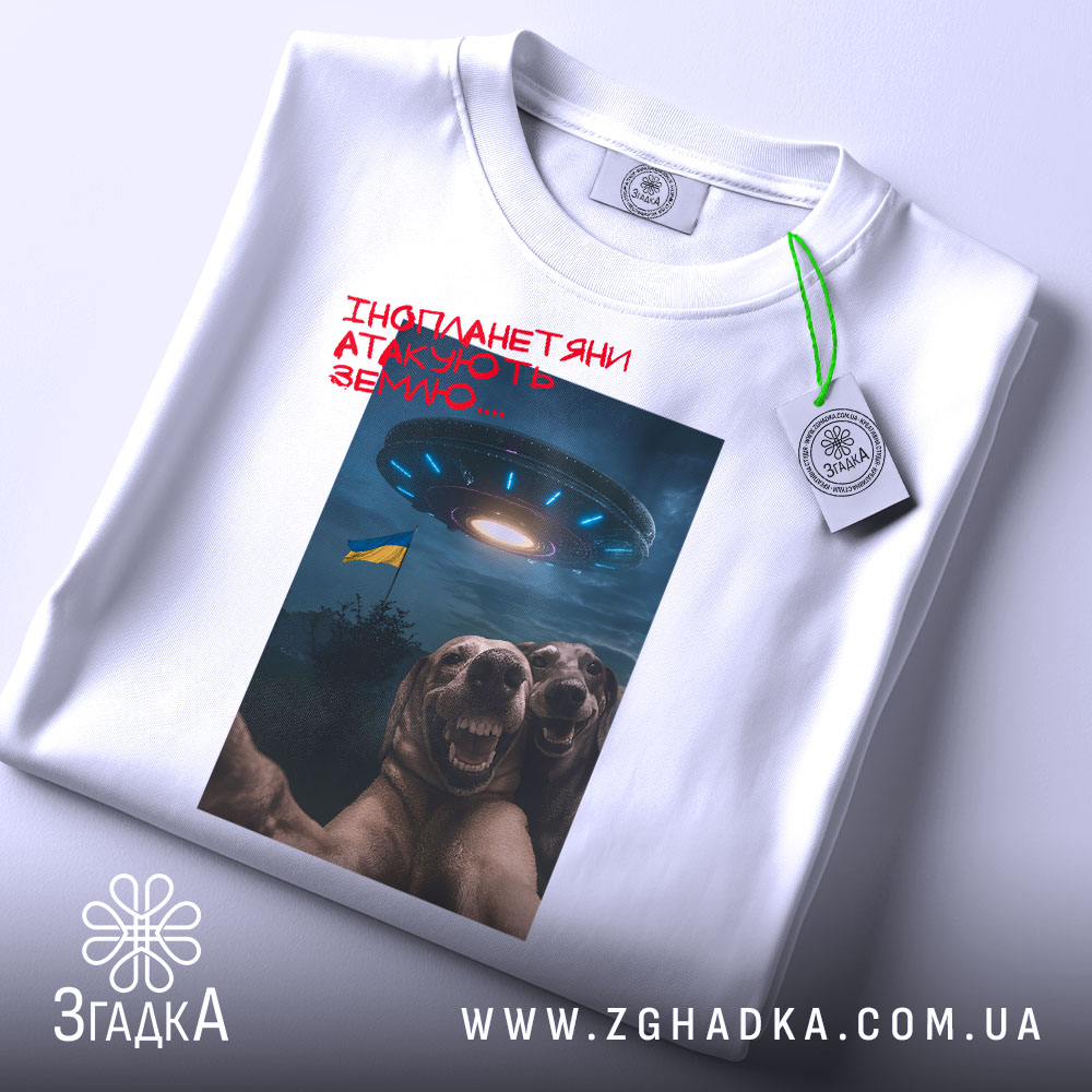 Футболка інопланетяни атакують Землю, біла футболка на столі, ракурс зверху, фон світлий, принт з двома собаками, що сміються, та космічним кораблем на фоні з українським прапором, з етикеткою.
