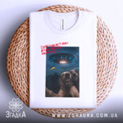 Футболка інопланетяни атакують Землю, біла футболка на плетеній кошику, ракурс зверху, фон натуральний, принт з двома собаками, що сміються, та космічним кораблем на фоні з українським прапором.