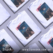 Футболка інопланетяни атакують Землю, біла футболка на манекені, ракурс спереду, фон нейтральний, принт з двома собаками, що сміються, та космічним кораблем на фоні з українським прапором.