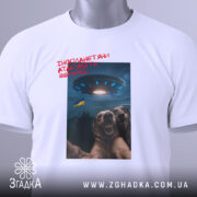 Футболка інопланетяни атакують Землю, біла футболка на столі, ракурс зверху, фон світлий, принт з двома собаками, що сміються, та космічним кораблем на фоні з українським прапором, з етикеткою.