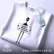 Футболка інопланетянин, біла, зображена зверху на плоскості, фон світло-сірий, принт чорного кольору з інопланетянином, що тримає табличку з написом 'I DON'T BELIEVE IN HUMANS', на футболці є етикетка з логотипом.