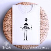 Футболка інопланетянин, біла, зображена зверху на плоскій поверхні, фон світло-сірий, принт чорного кольору з інопланетянином, що тримає табличку з написом 'I DON'T BELIEVE IN HUMANS', футболка лежить на плетеній основі.
