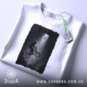 Футболка інопланетна зустріч, біла, зображена на столі, фон світло-сірий, принт з хлопчиком на велосипеді під космічним кораблем, з етикеткою зеленого кольору.