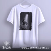 Футболка інопланетна зустріч, біла, підвішена на вішалці, фон світло-сірий, принт з хлопчиком на велосипеді, що летить під космічним кораблем, в темних тонах.