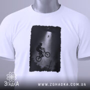 Футболка інопланетна зустріч, біла, зображена на столі, фон світло-сірий, принт з хлопчиком на велосипеді під космічним кораблем, з етикеткою зеленого кольору.