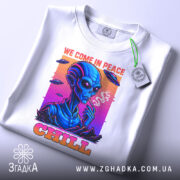 Футболка інопланетний чіл, біла, складена на столі, фон світло-сірий, принт з інопланетянином, який курить, кольори принта яскраві: синій, рожевий, оранжевий, текст 'WE COME IN PEACE' і 'CHILL', на футболці є етикетка з зеленим шнурком.