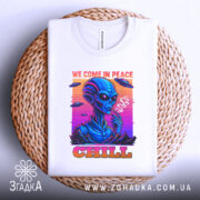 Футболка інопланетний чіл, біла, складена на плетеній кошику, фон світло-сірий, принт з інопланетянином, який курить, кольори принта яскраві: синій, рожевий, оранжевий, текст 'WE COME IN PEACE' і 'CHILL', видно етикетку на горловині.