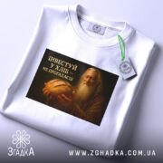 Футболка інвестуй у хліб, складена на поверхні, фон білий, принт зображує чоловіка з бородою, який тримає хліб, текст на принті чорний з жовтим, на футболці є етикетка з зеленим шнурком.