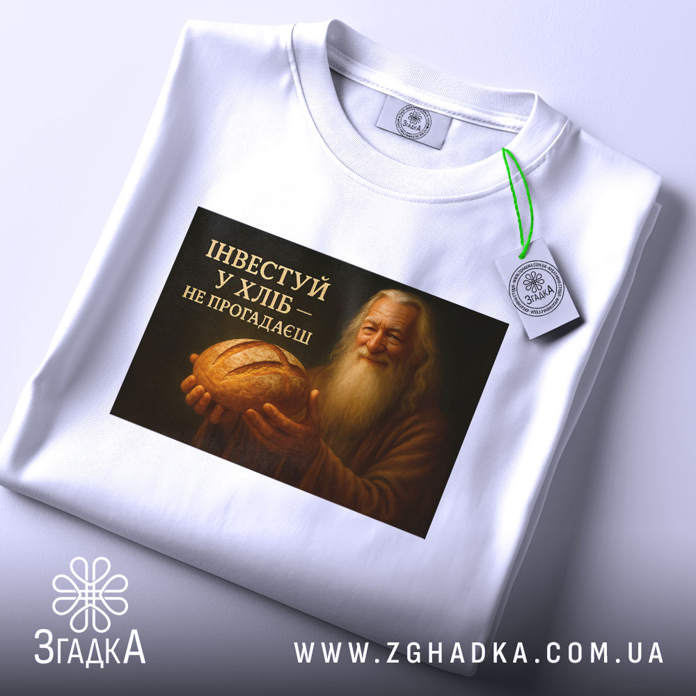 Футболка інвестуй у хліб, складена на поверхні, фон білий, принт зображує чоловіка з бородою, який тримає хліб, текст на принті чорний з жовтим, на футболці є етикетка з зеленим шнурком.