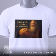 Футболка інвестуй у хліб, складена на поверхні, фон білий, принт зображує чоловіка з бородою, який тримає хліб, текст на принті чорний з жовтим, на футболці є етикетка з зеленим шнурком.