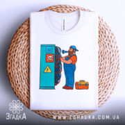 Футболка інженер електрик, біла, зображення зверху, фон світло-сірий, принт з чоловіком у синьому комбінезоні, який працює з електричними проводами, футболка лежить на плетеному кошику.