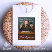 Футболка Іван Франко, біла, складена на плетеній кошику, фон світлий, принт з портретом Івана Франка, текст "Лиш боротись – значить жить!" на зеленому фоні, чоловік у костюмі з бородою, тримає телефон і показує знак миру.