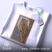 Футболка із совою білого кольору, складена на поверхні, ракурс зверху, фон світло-сірий, принт сови на гілці з деталями лісу, прикріплена етикетка з зеленим шнурком.