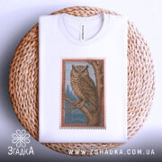 Футболка із совою білого кольору, складена на плетеній кошику, ракурс зверху, фон світло-сірий, принт сови на гілці з деталями лісу, видима етикетка.