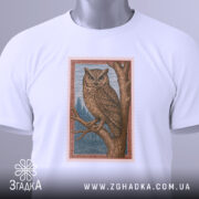 Футболка із совою білого кольору, складена на поверхні, ракурс зверху, фон світло-сірий, принт сови на гілці з деталями лісу, прикріплена етикетка з зеленим шнурком.