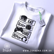 Футболка їж спи білого кольору, зображена зверху, на ній чорний принт з написами 'EAT', 'SLEEP', 'GAME', 'REPEAT' та ілюстраціями, на футболці є зелений ярлик, фон світлий.