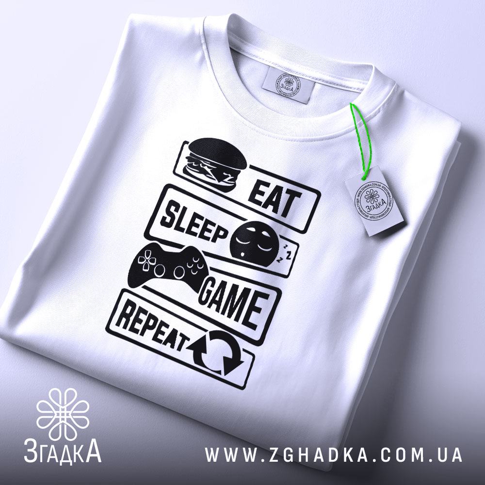 Футболка їж спи білого кольору, зображена зверху, на ній чорний принт з написами 'EAT', 'SLEEP', 'GAME', 'REPEAT' та ілюстраціями, на футболці є зелений ярлик, фон світлий.