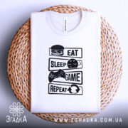 Футболка їж спи білого кольору, зображена зверху на плетеній основі, фон світлий, принт чорного кольору з написами 'EAT', 'SLEEP', 'GAME', 'REPEAT' та ілюстраціями, видна етикетка на комірці.