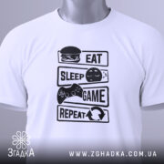 Футболка їж спи білого кольору, зображена зверху, на ній чорний принт з написами 'EAT', 'SLEEP', 'GAME', 'REPEAT' та ілюстраціями, на футболці є зелений ярлик, фон світлий.