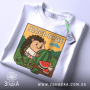 Футболка їжачок оптиміст. Біла футболка, складена на поверхні, з принтом їжачка, що тримає кавун. На фоні видно зелені пагорби та блакитне небо, принт яскравий і детальний, з елементами природи.