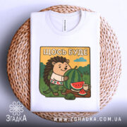 Футболка їжачок оптиміст. Біла футболка, складена на плетеній кошику, з принтом їжачка, що тримає кавун. Принт яскравий, фон з зеленими пагорбами та блакитним небом, деталі принта чіткі.